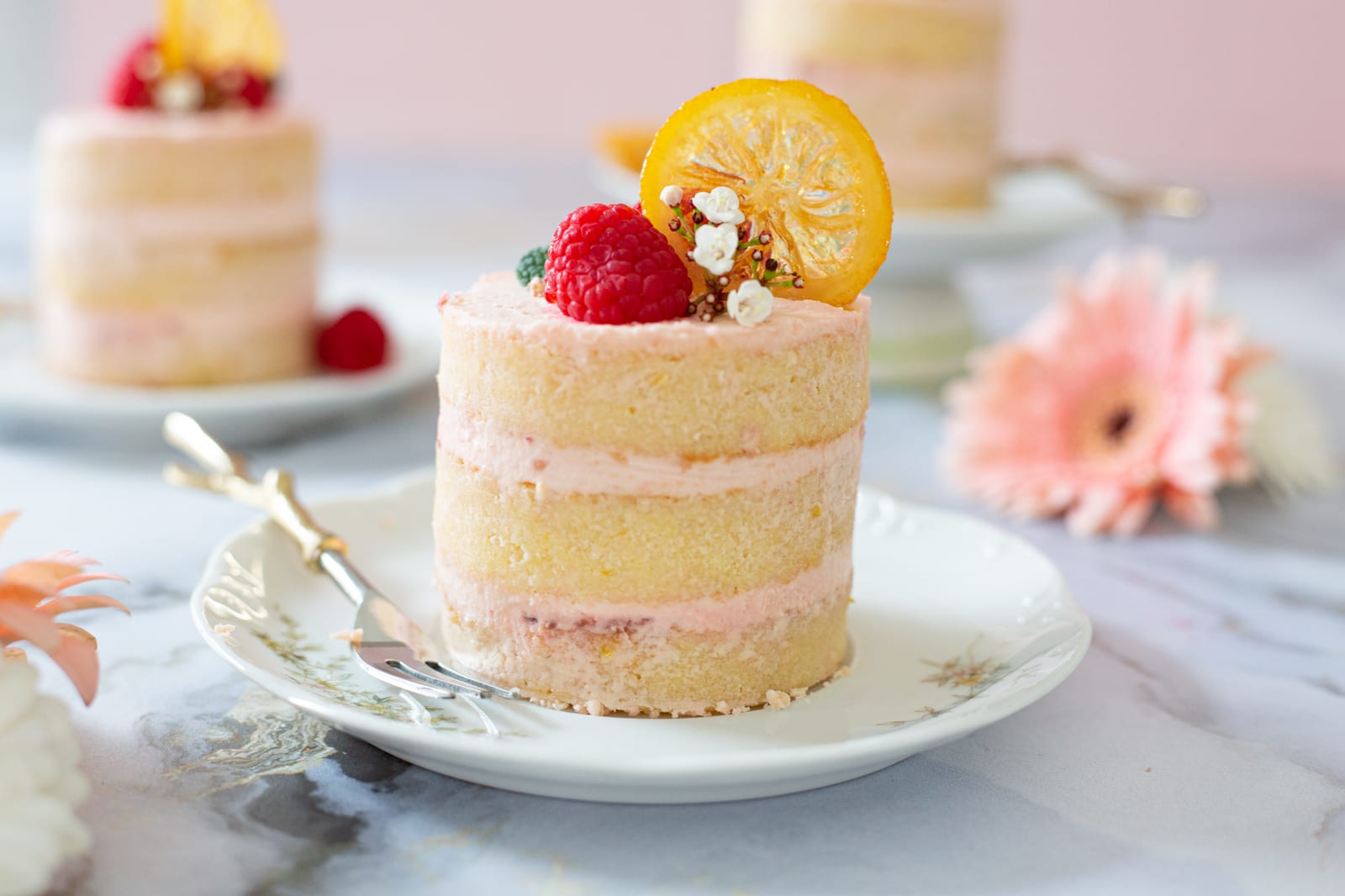 A raspberry and lemon mini cake on a plate with another behind, pink gerberas around