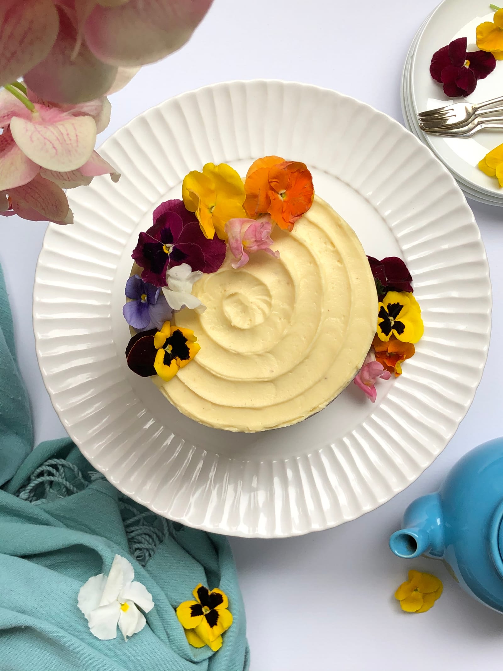 A whole carrot cake decorated with edible pansies on a white fluted plate, a teapot and pink flowers beside it, from above