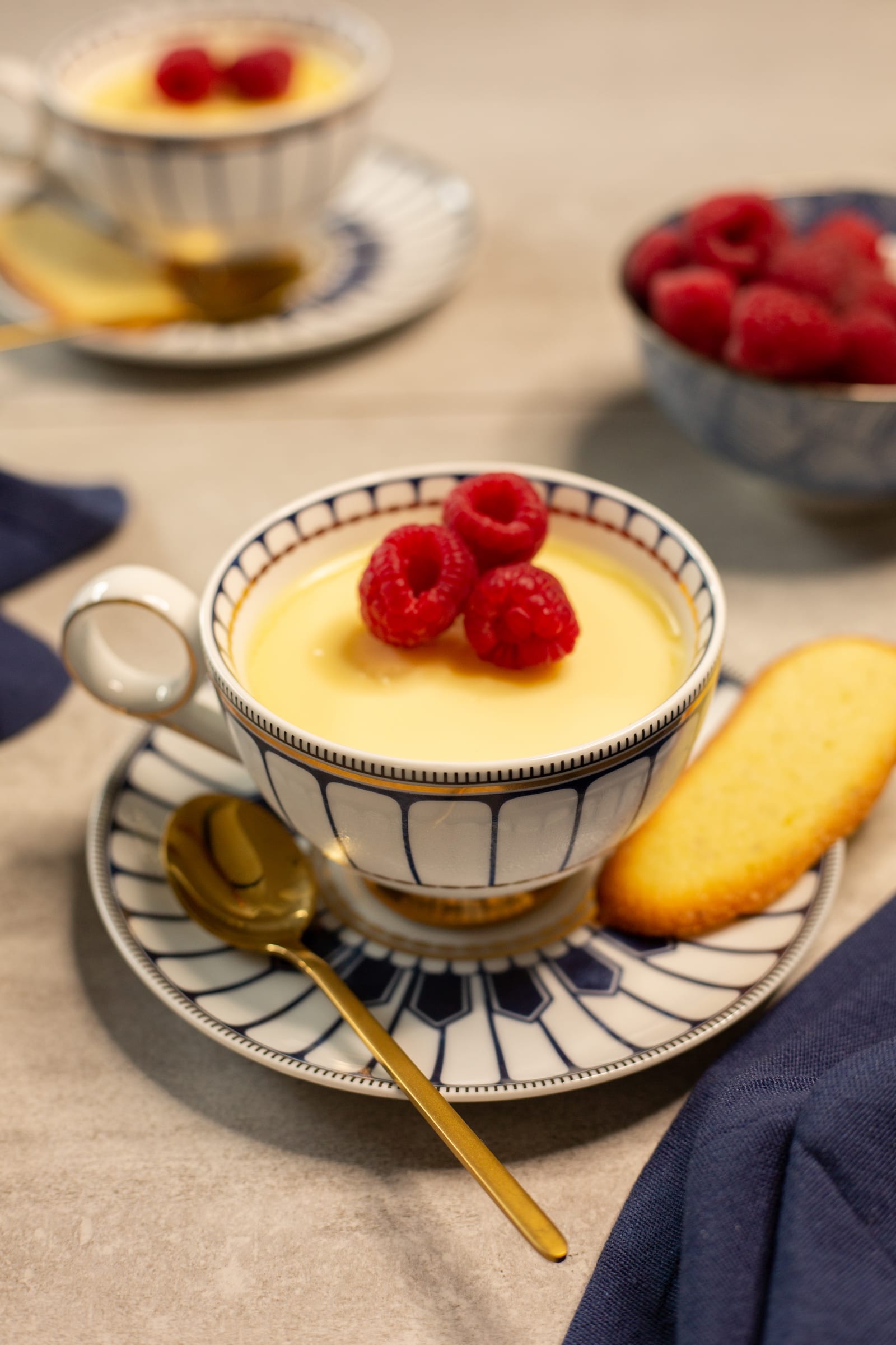 A lemon posset in a patterned teacup topped with raspberries, with a cat's tongue biscuit and a gold spoon