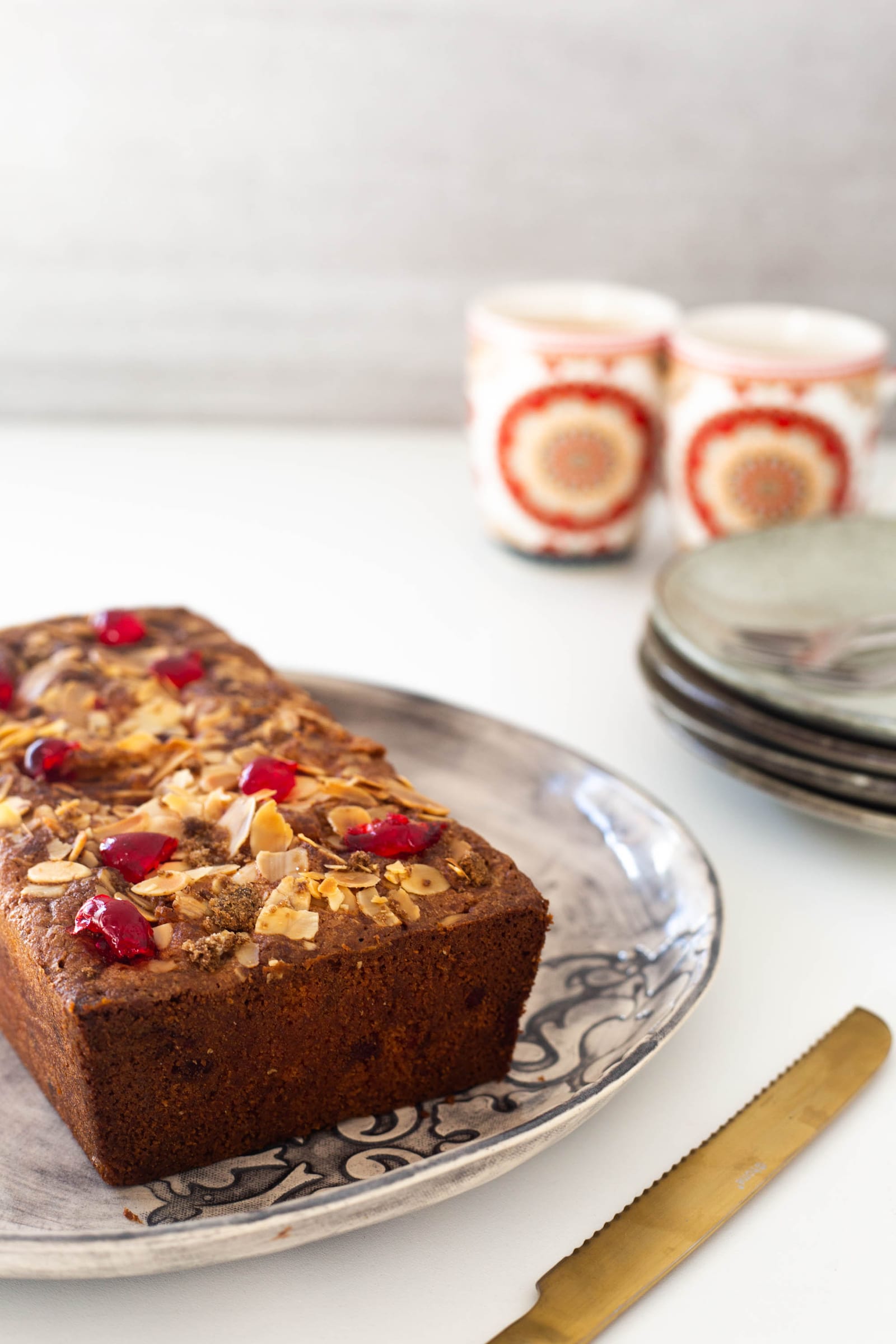 A gluten-free cherry and almond loaf on a patterned plate beside mugs and stacked plates