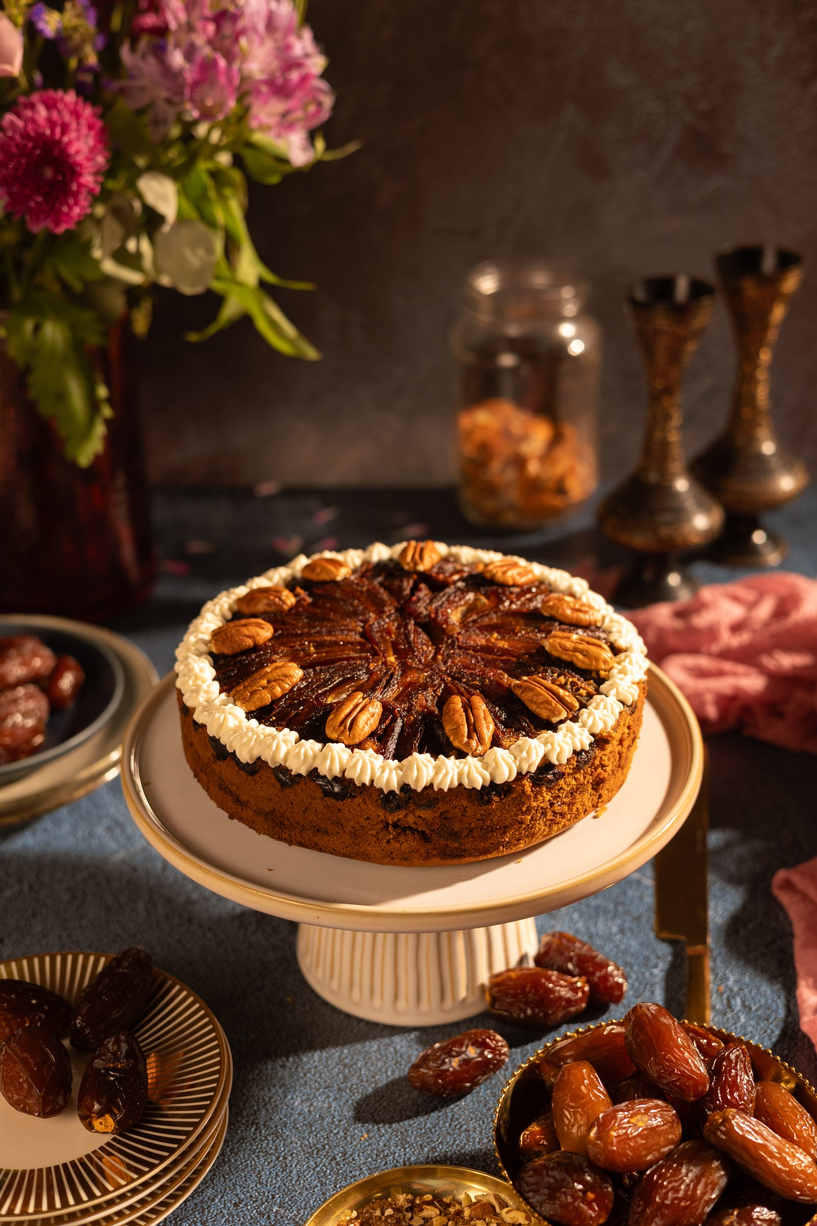 A sticky date and pecan cake on a cake stand, with flowers and dates