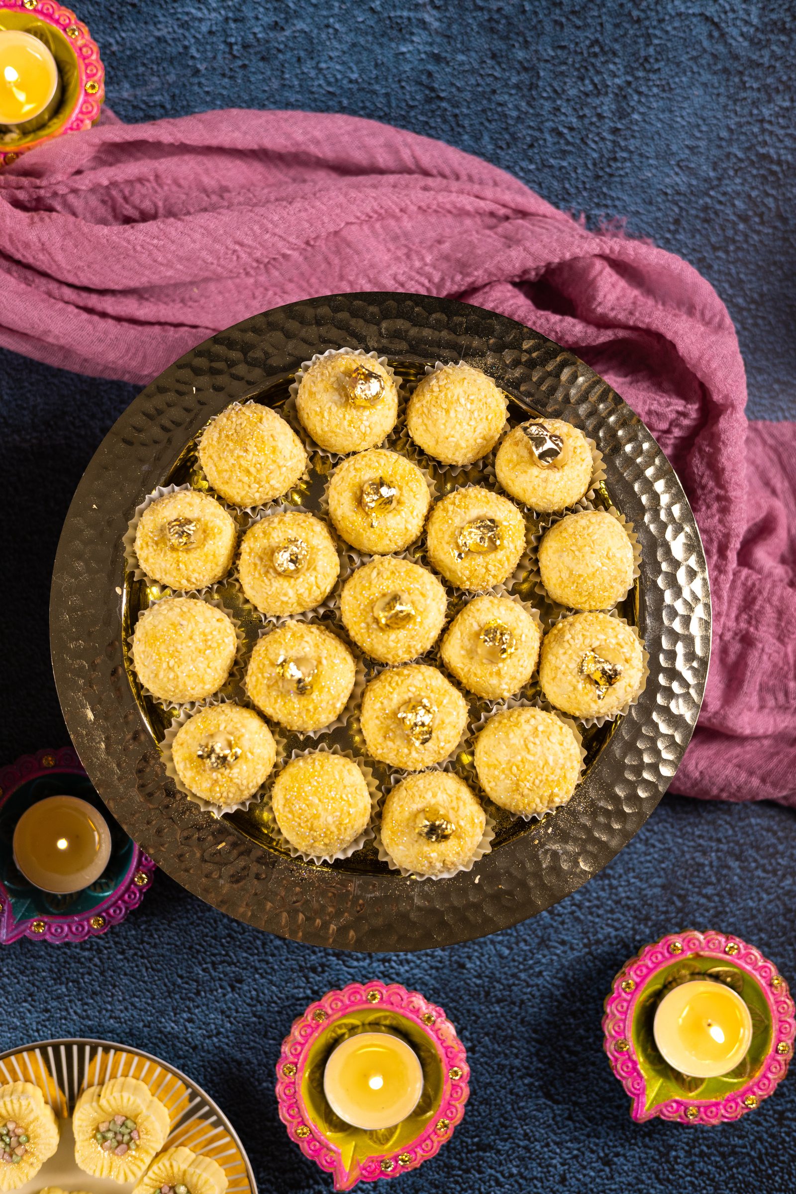 A hammered copper platter of coconut Simply Wonderfuls on a blue cloth surrounded by diyas, from above