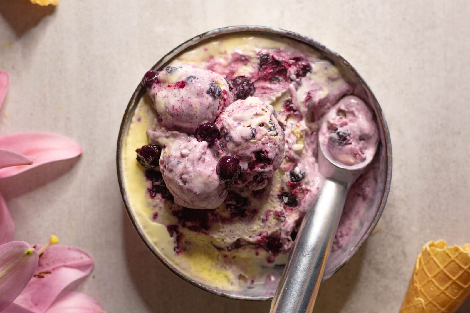 A tin of scooped blueberry swirl ice cream with a metal scoop, a cone and pink lilies, from above