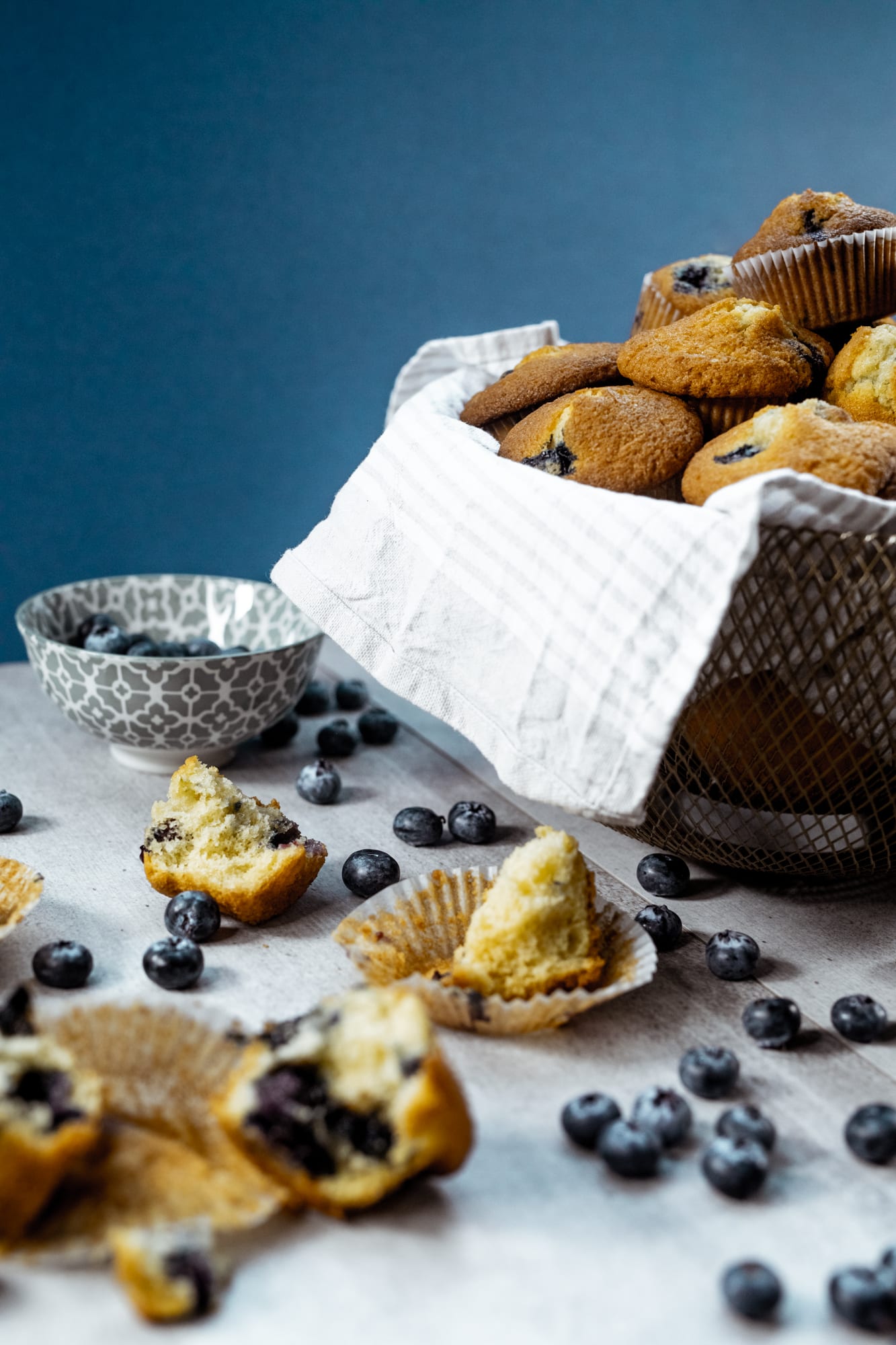 Broken-open one bowl blueberry muffins beside the basket and a bowl of blueberries