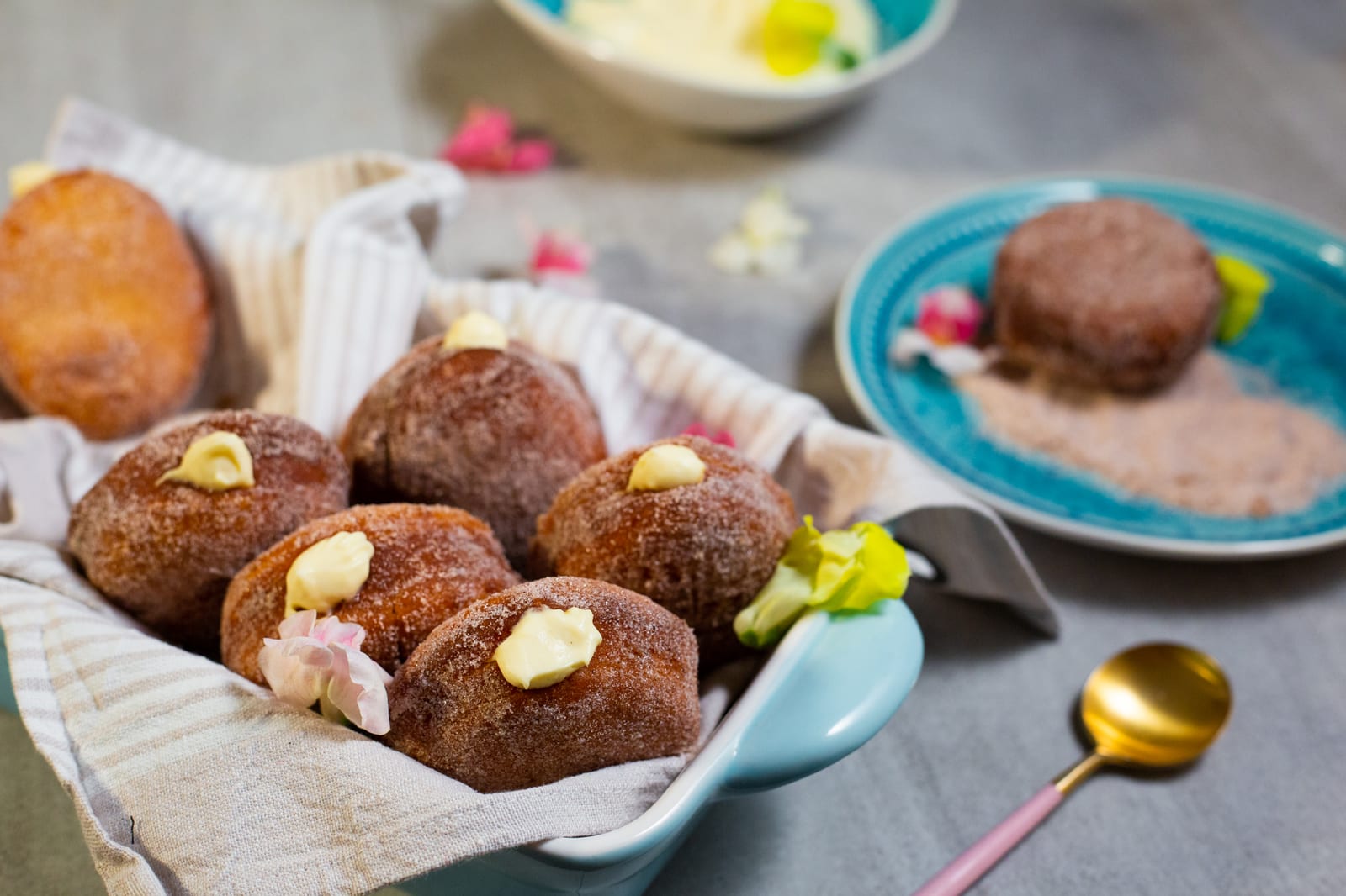 Doughnuts filled with milk tart mousse in a cloth-lined dish and on a teal plate of cinnamon sugar, a bowl of mousse beside them