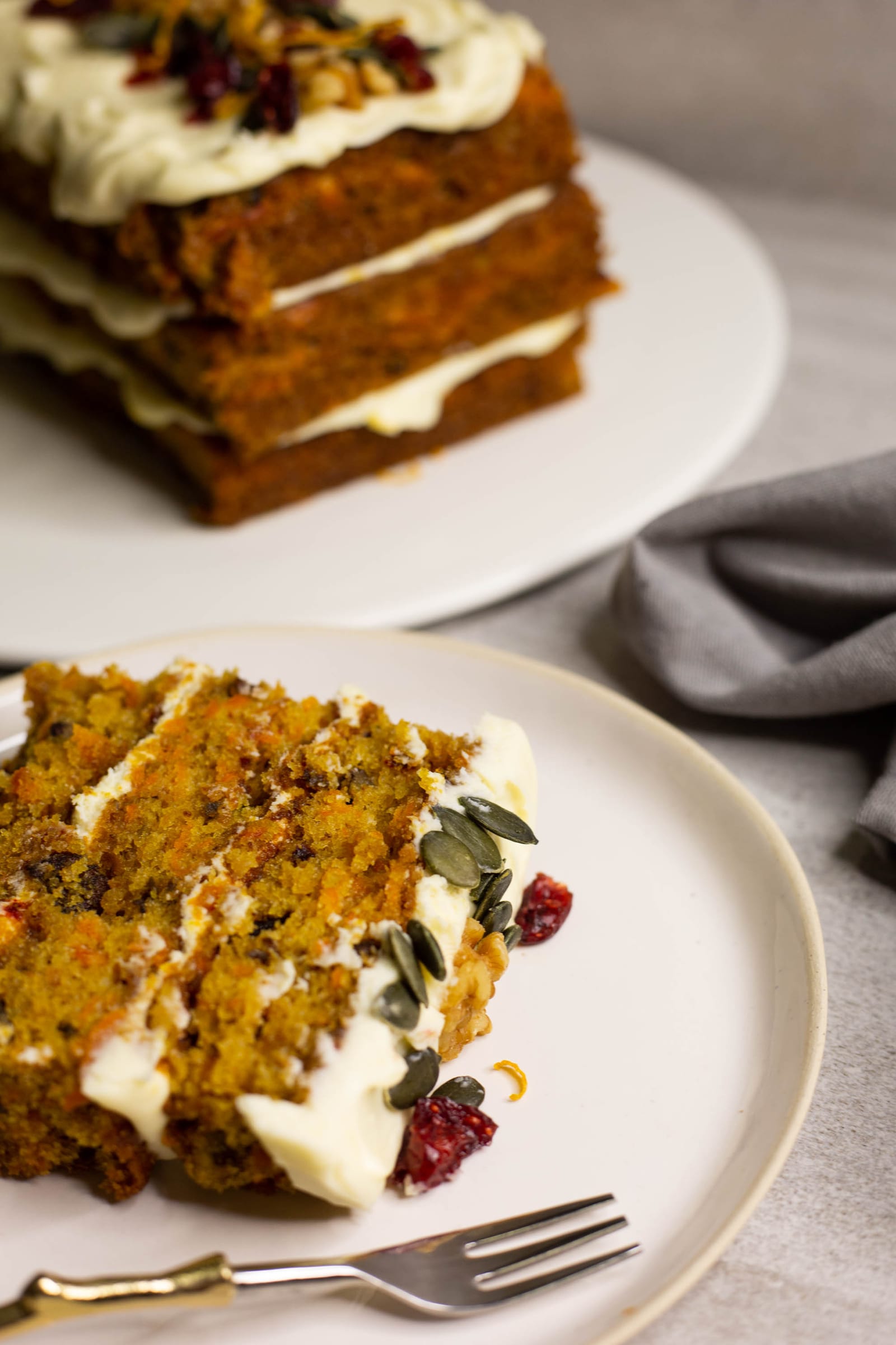 A slice of carrot cake with orange cream cheese icing on a plate, with the cake behind