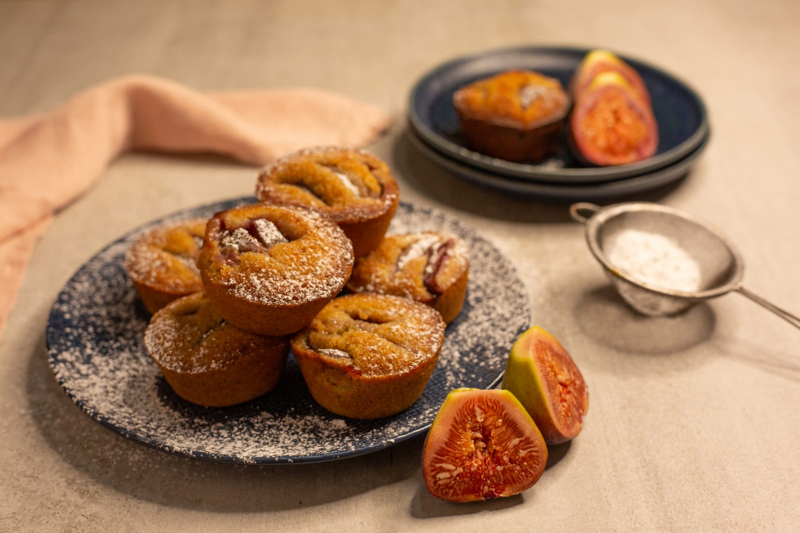 A pile of fig and hazelnut friands on a navy plate with halved figs and a sieve of icing sugar