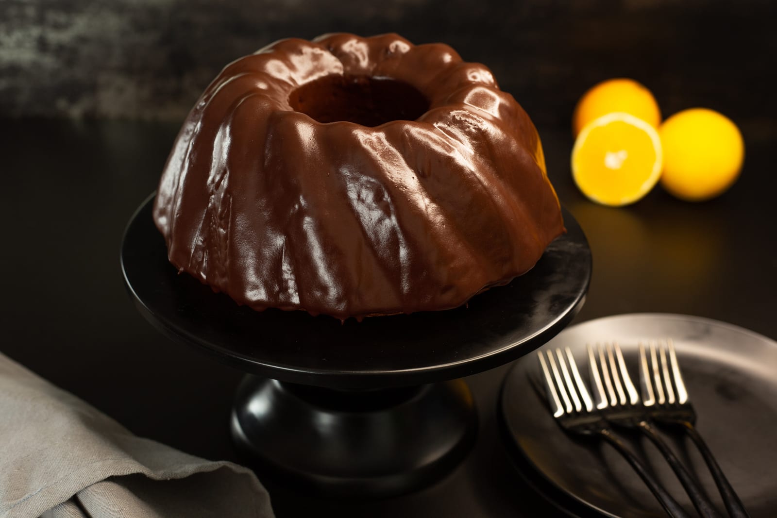 An orange and olive oil bundt with a glossy dark chocolate glaze on a black cake stand, with orange halves beside it