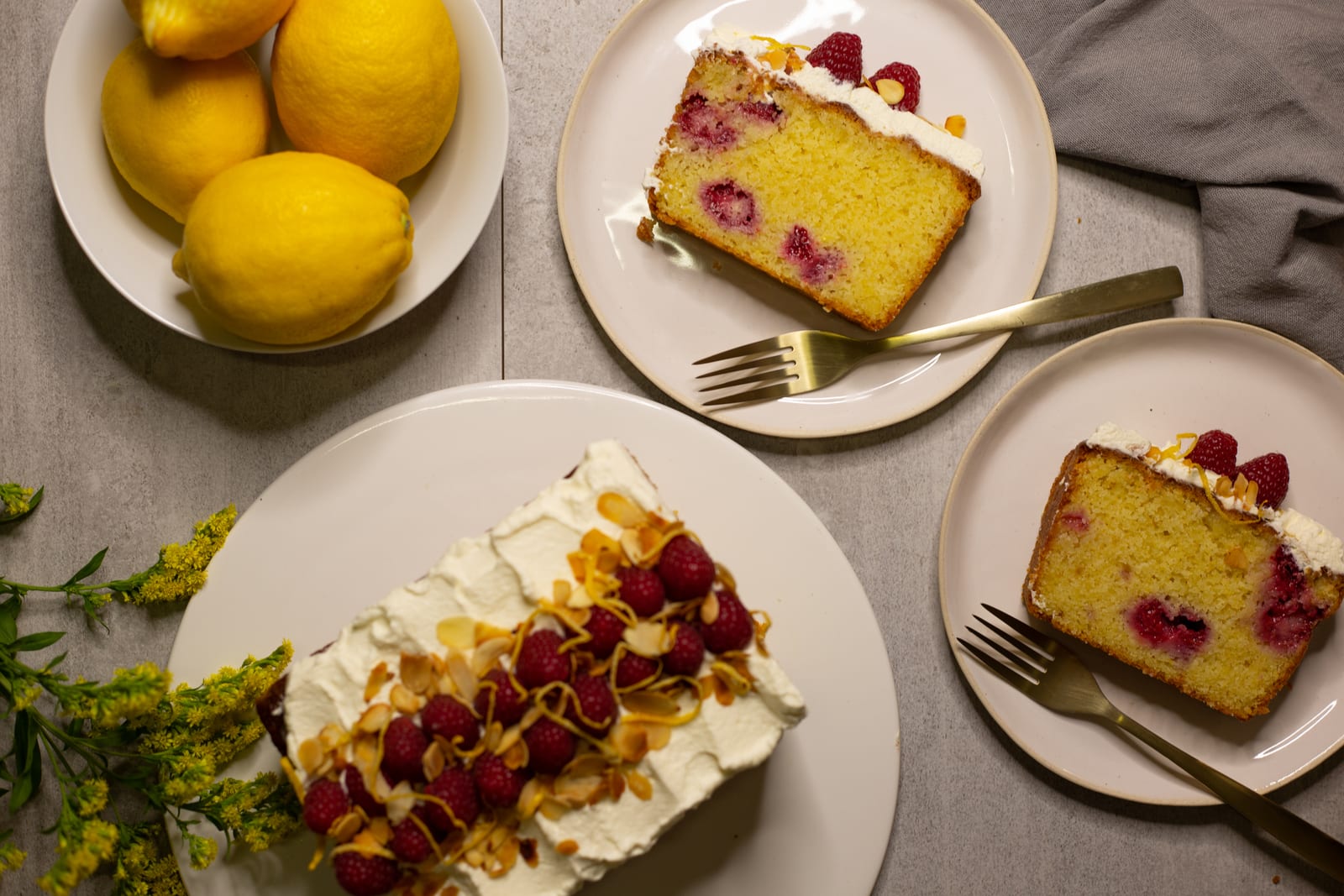 A gluten-free almond, raspberry and lemon loaf with slices on small plates and gold forks, lemons beside it, from above