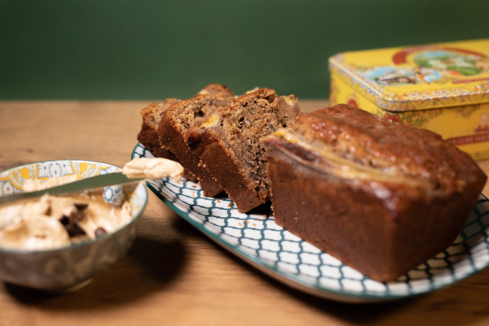 A banana and pecan nut bread partly sliced on a blue and white dish, with a bowl of whipped butter and a yellow tin