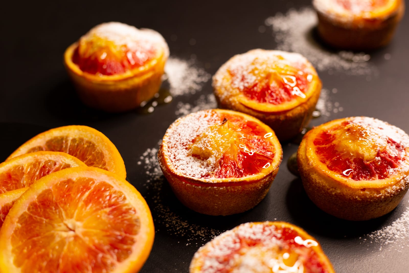 A row of almond, semolina and blood orange syrup cakes dusted with icing sugar among blood orange slices