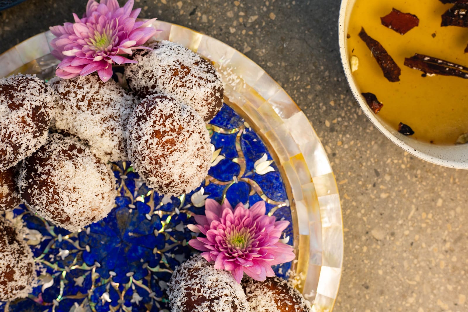 Cape Malay koesisters dusted in coconut on a blue mosaic plate, with pink flowers and a bowl of spiced syrup