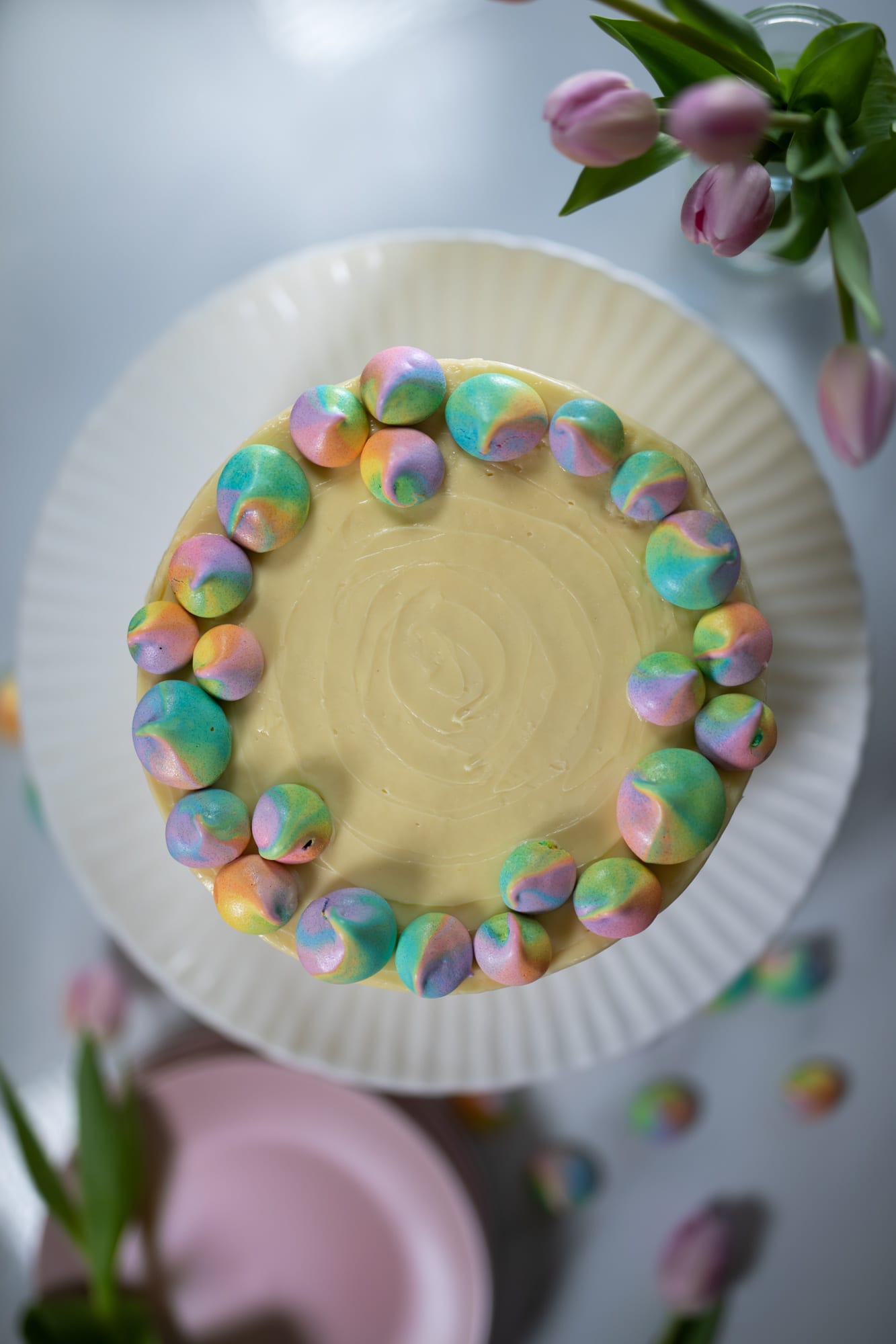 A whole pastel rainbow cake ringed with meringue kisses beside tulips and a pink plate, from above