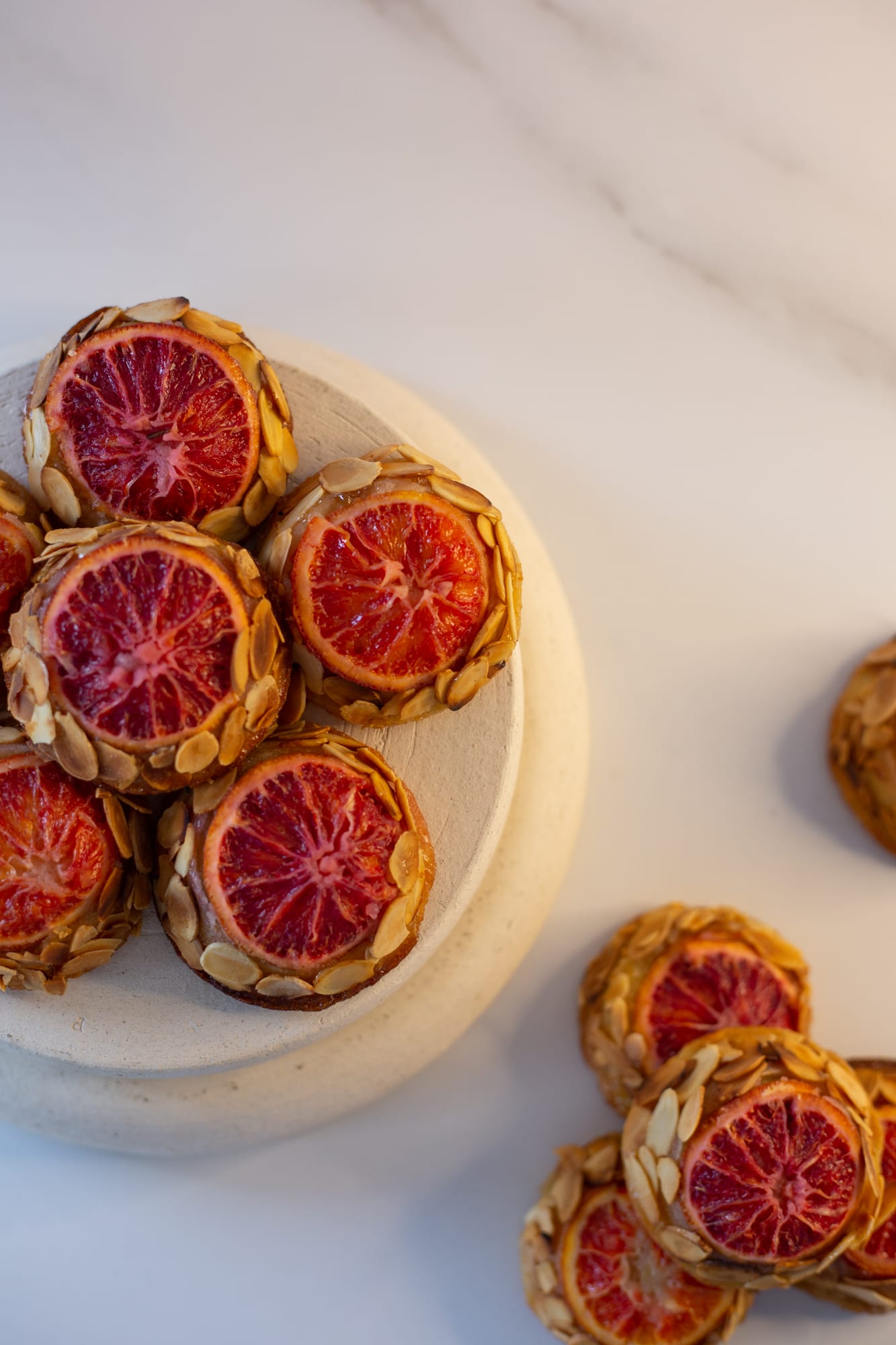 Blood orange and almond tea cakes piled on a two-tier stone riser and scattered on marble, from above