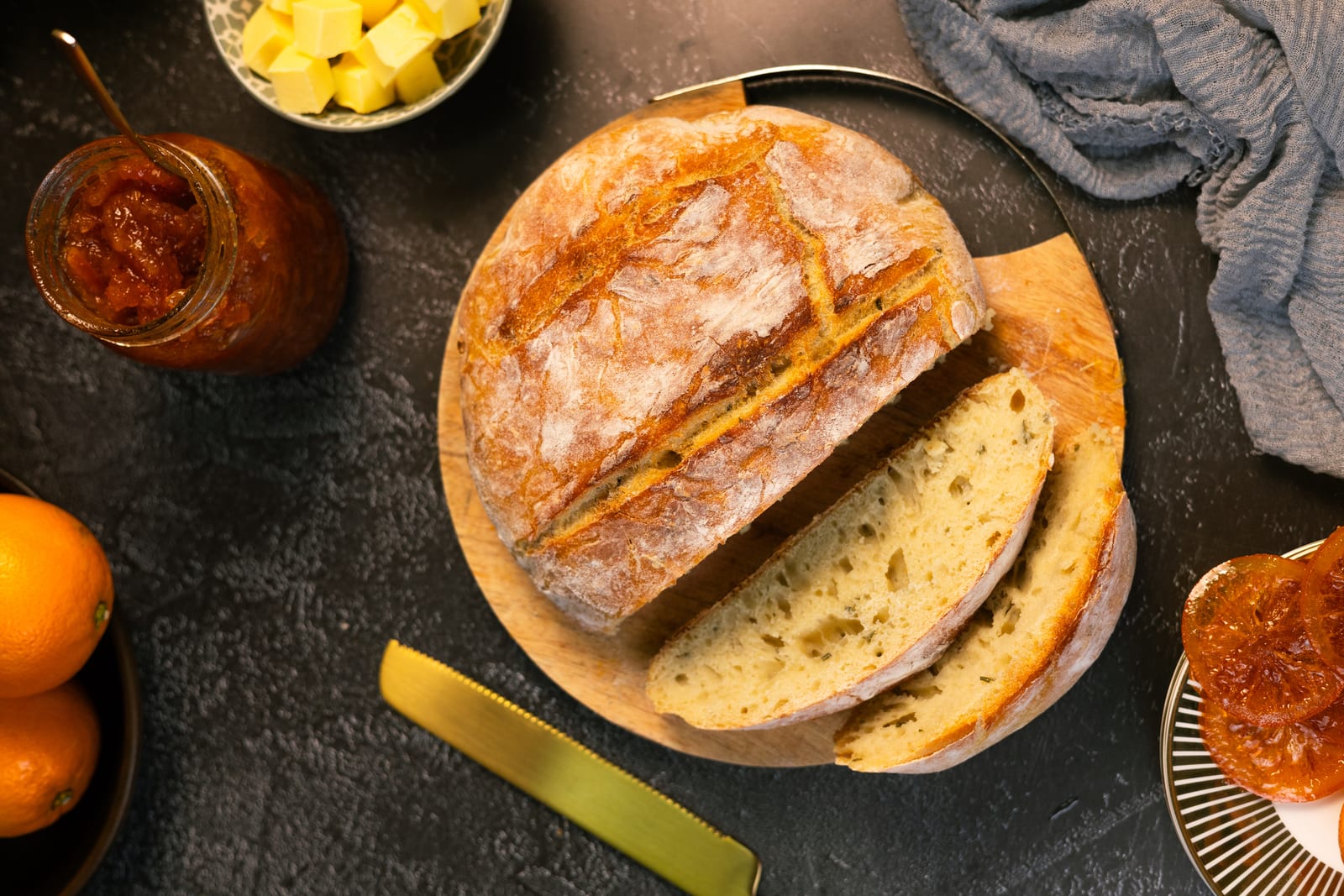 A partly sliced no-knead rosemary loaf on a wooden board with marmalade, butter and oranges, from above
