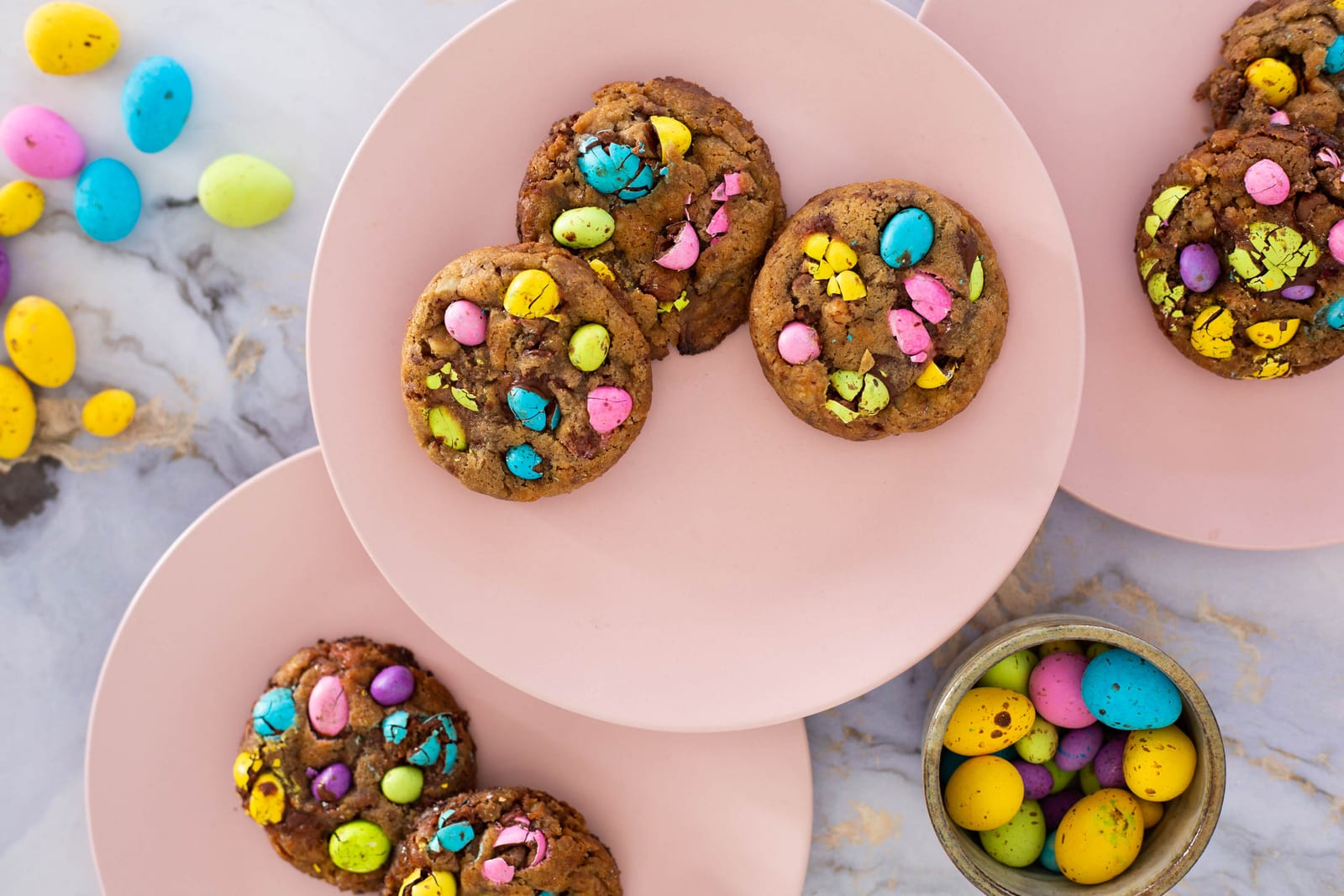 Speckled egg choc chunk cookies on pink plates beside a brass bowl of speckled eggs, from above