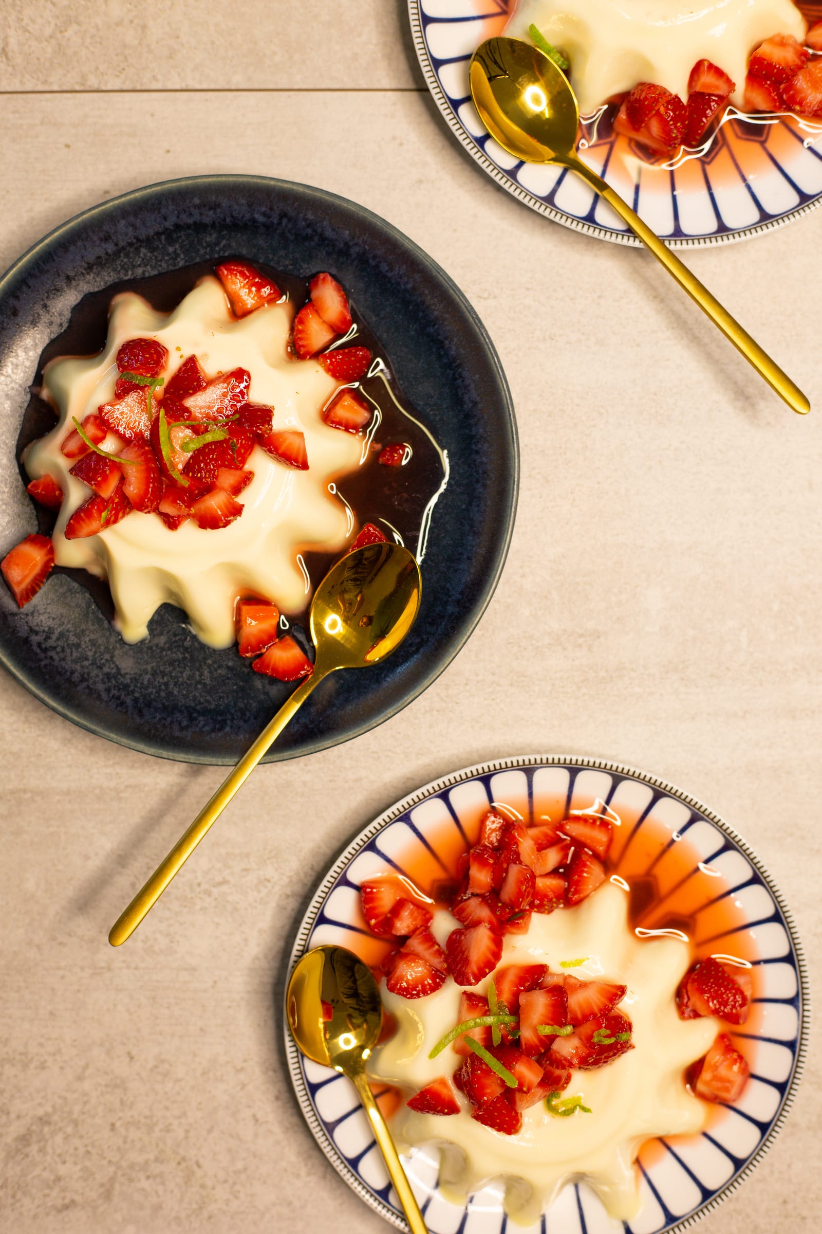 A lime panna cotta with strawberries on a dark blue plate with gold spoons, from above