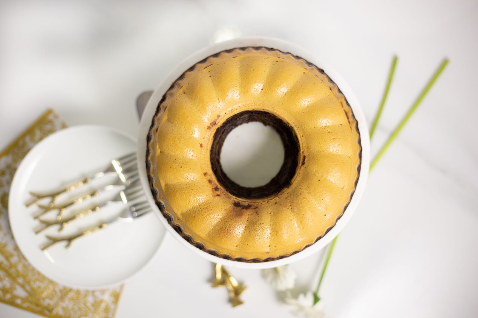A whole caramel white chocoflan on a cake stand beside plates, gold forks and white gerberas, from above