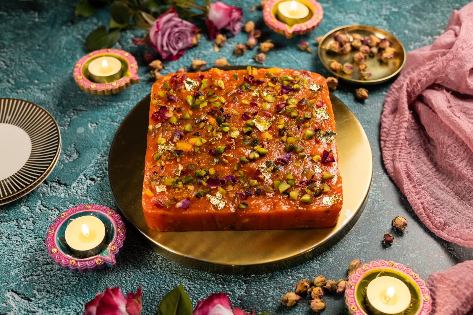 A whole slab of halwa topped with pistachios, rose petals and gold leaf on a gold board, surrounded by diyas and roses