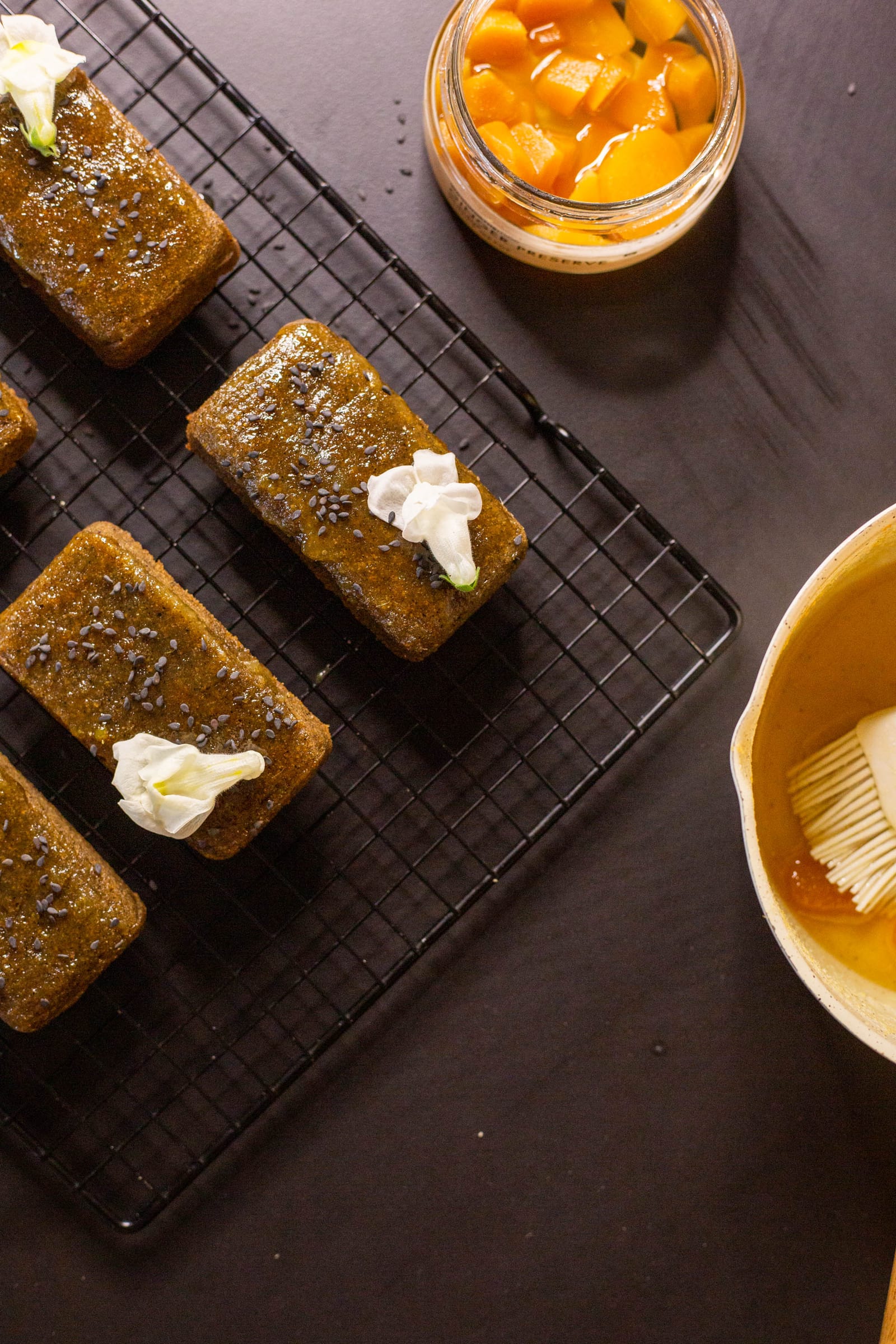 Black sesame and ginger financiers on a wire rack beside a jar of candied ginger, from above