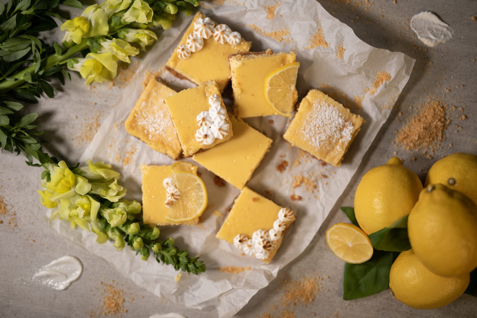 Sweet and tart lemon squares on parchment with green snapdragons and whole lemons, from above