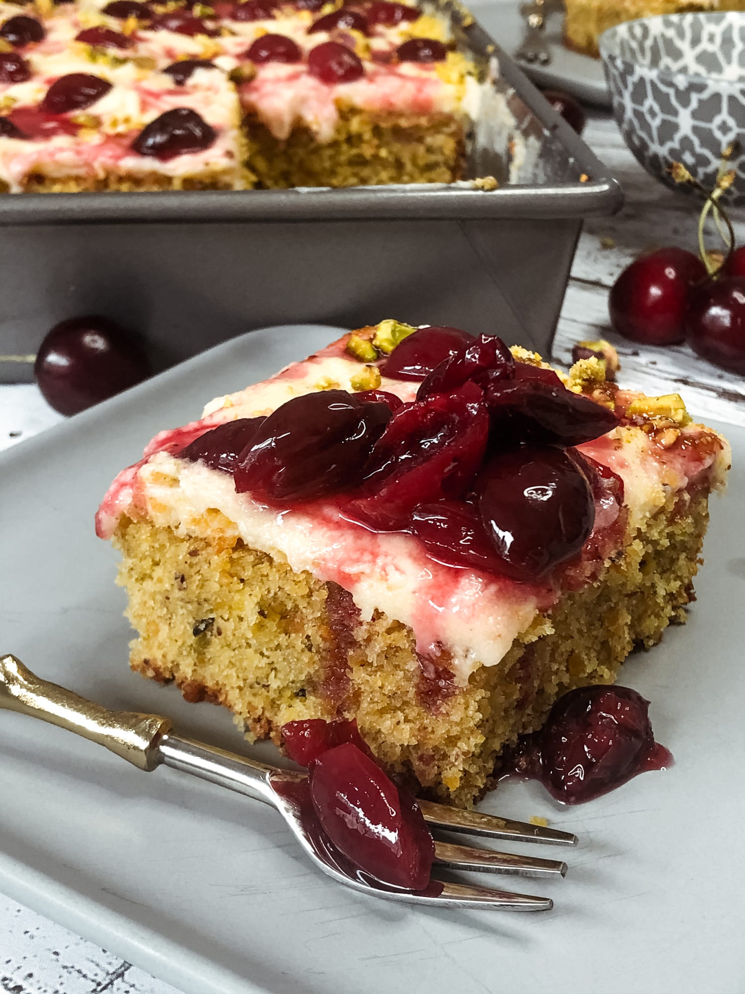 A square of pistachio and cherry sheet cake topped with cherries, the tin behind