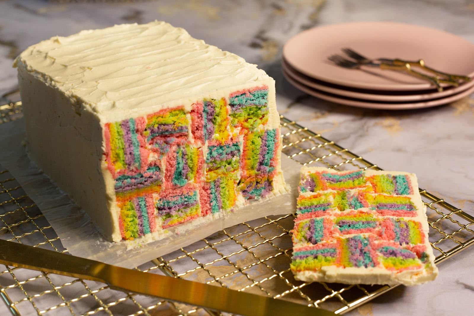 A cut rainbow coconut layer cake showing its woven rainbow squares, a slice and pink plates beside it