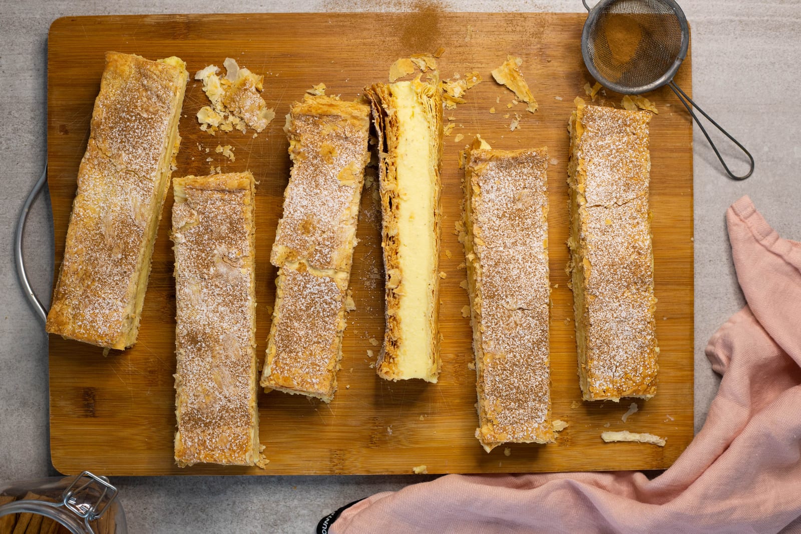 Milk tart slices cut into bars on a wooden board with a sieve of cinnamon sugar, from above