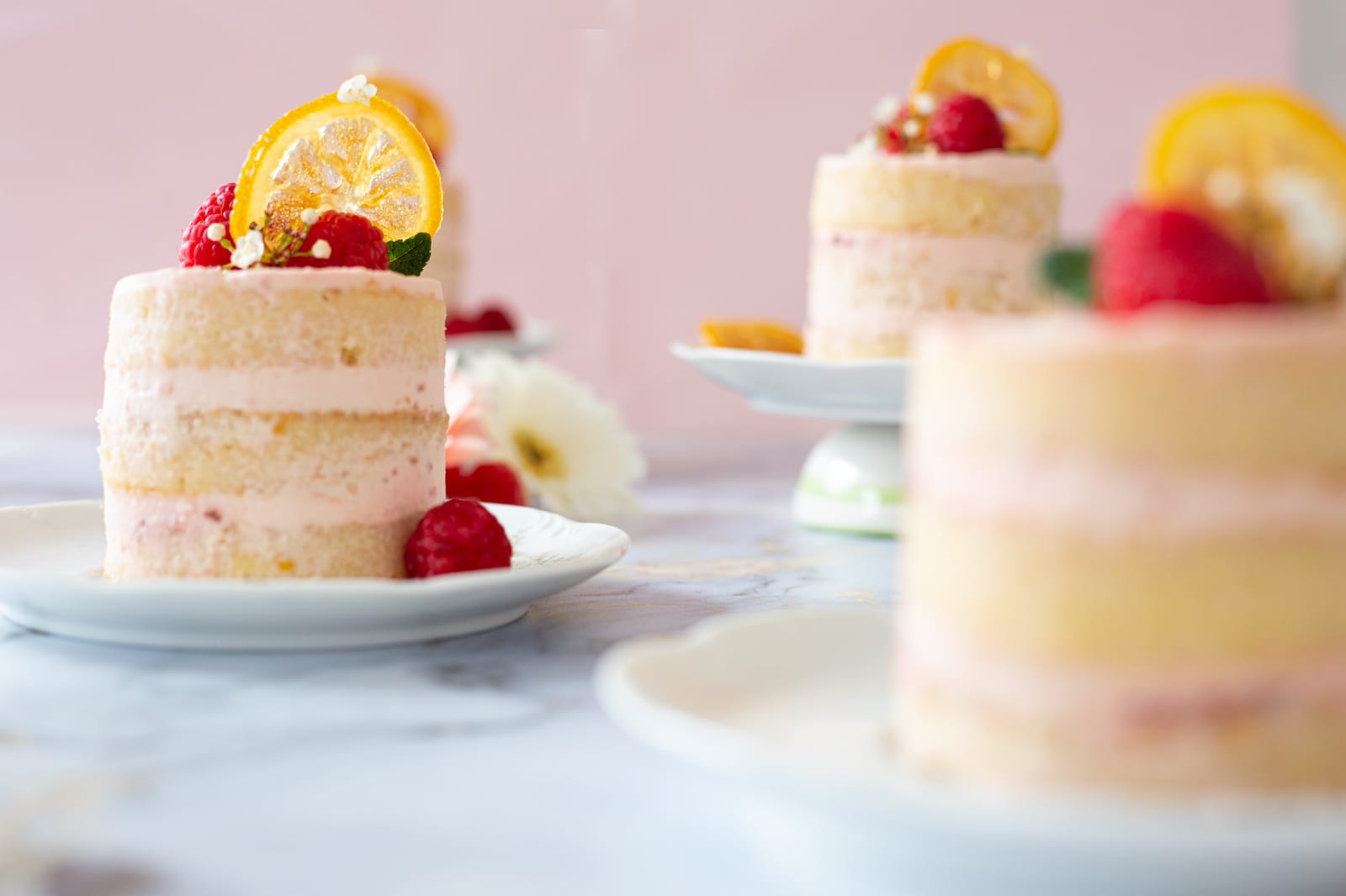 Three raspberry and lemon mini cakes topped with raspberries and candied lemon slices against a pink backdrop