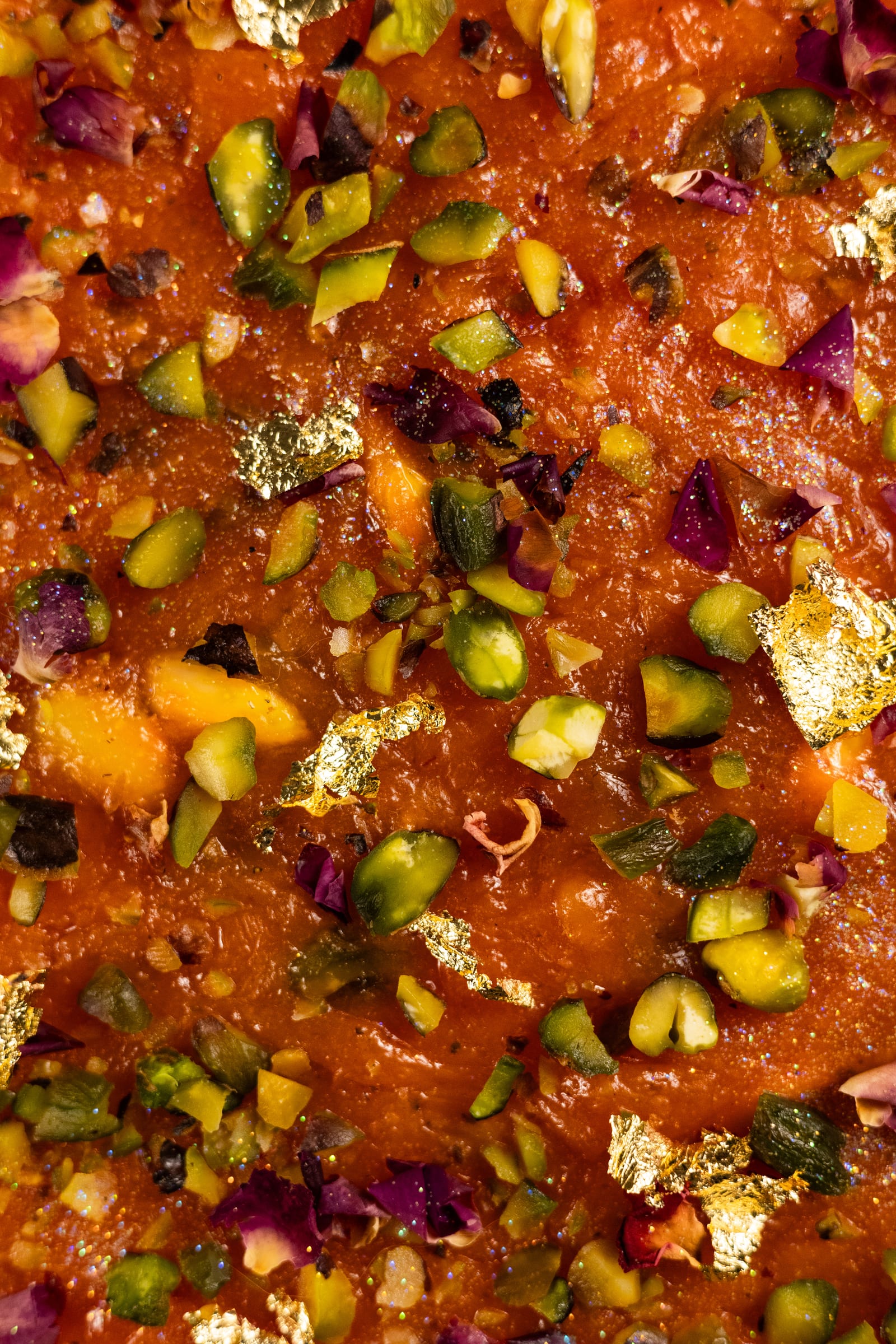 The glossy top of the halwa studded with pistachios, almonds, rose petals and gold leaf, close up