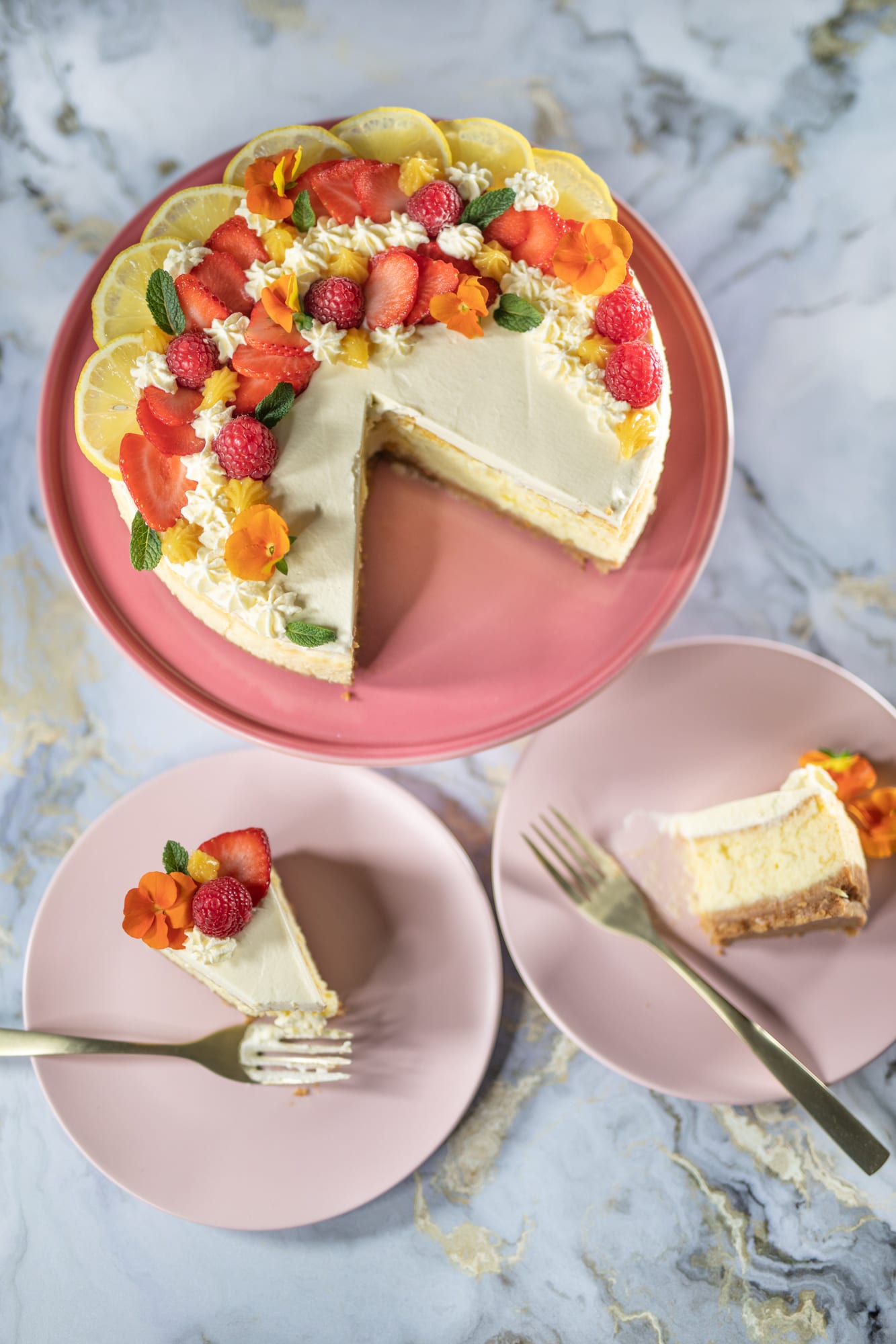 A cut creamy lemon cheesecake beside two slices on plates with gold forks, from above