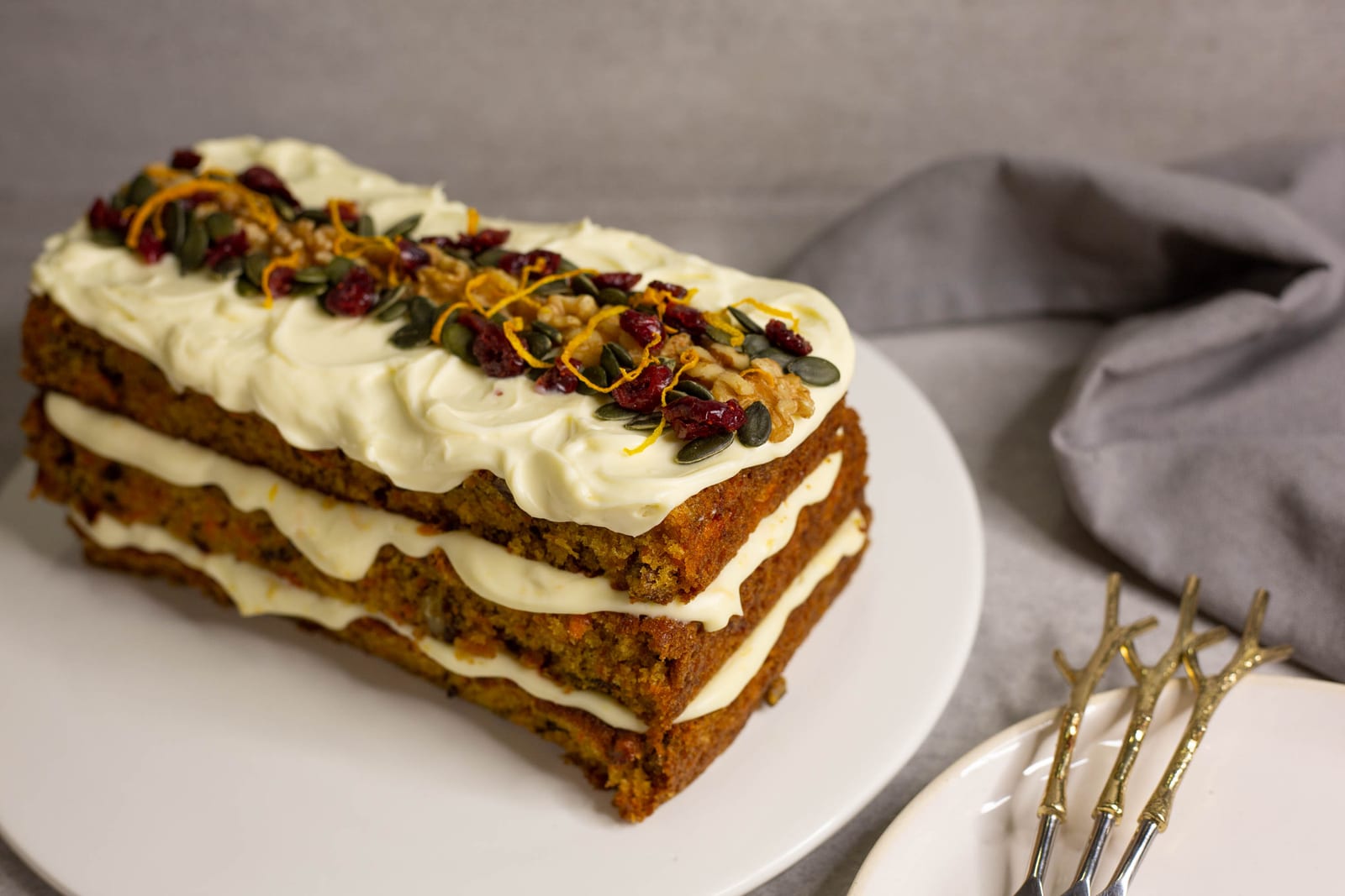 A three-layer carrot cake with orange cream cheese icing, topped with walnuts, cranberries and seeds, with gold forks alongside