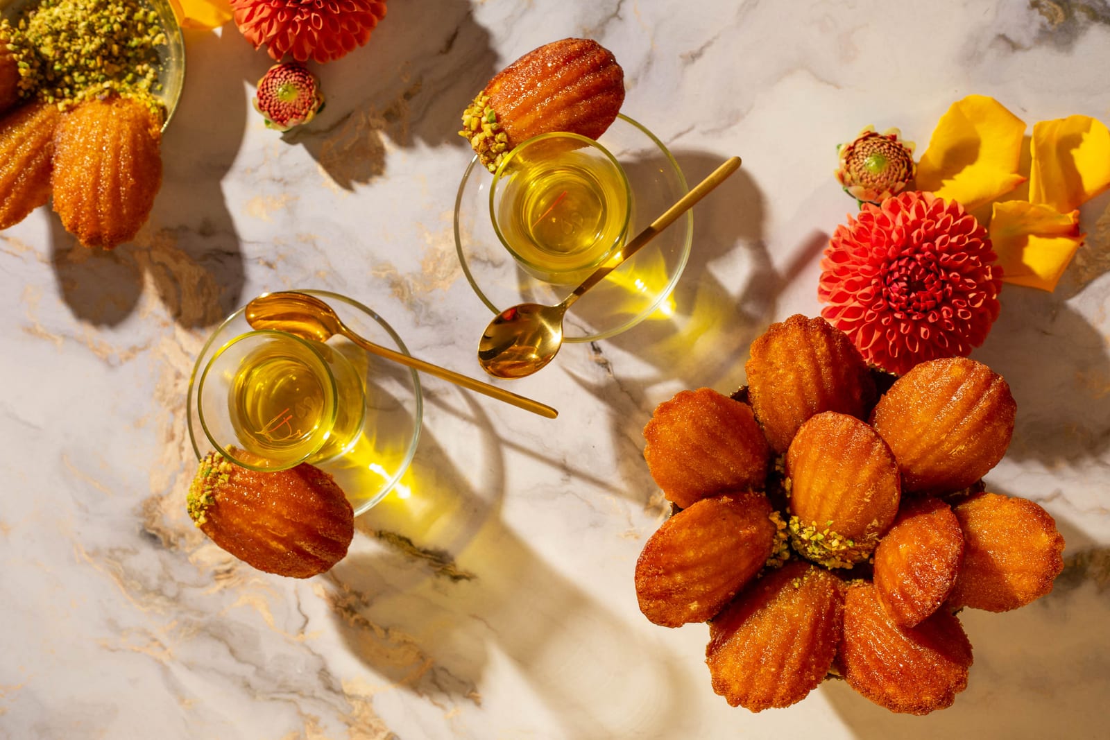 Orange blossom water madeleines arranged like a flower around crushed pistachios, beside tea glasses, from above