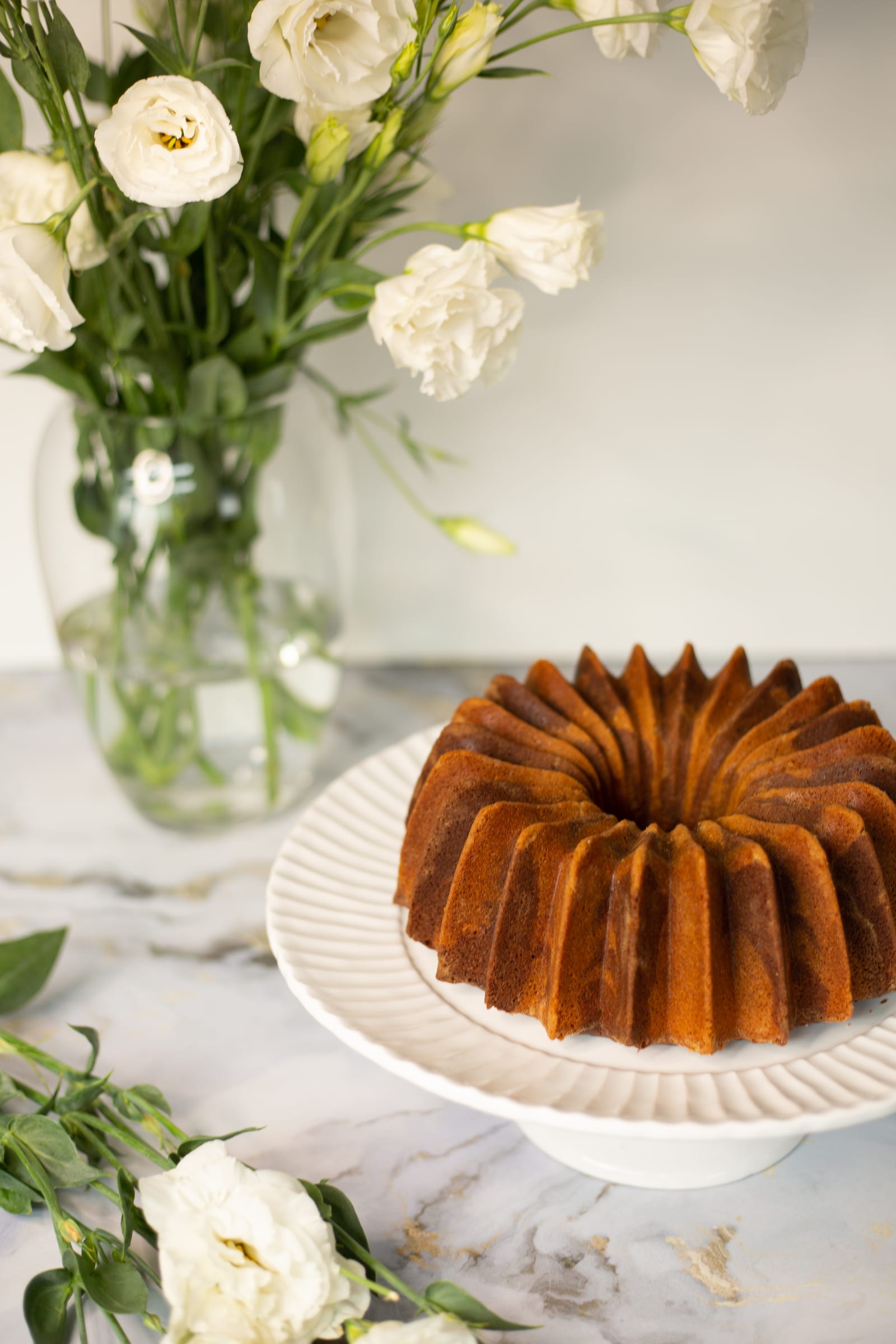 A whole marble pound cake baked in a fluted bundt tin, on a white cake stand beside white flowers