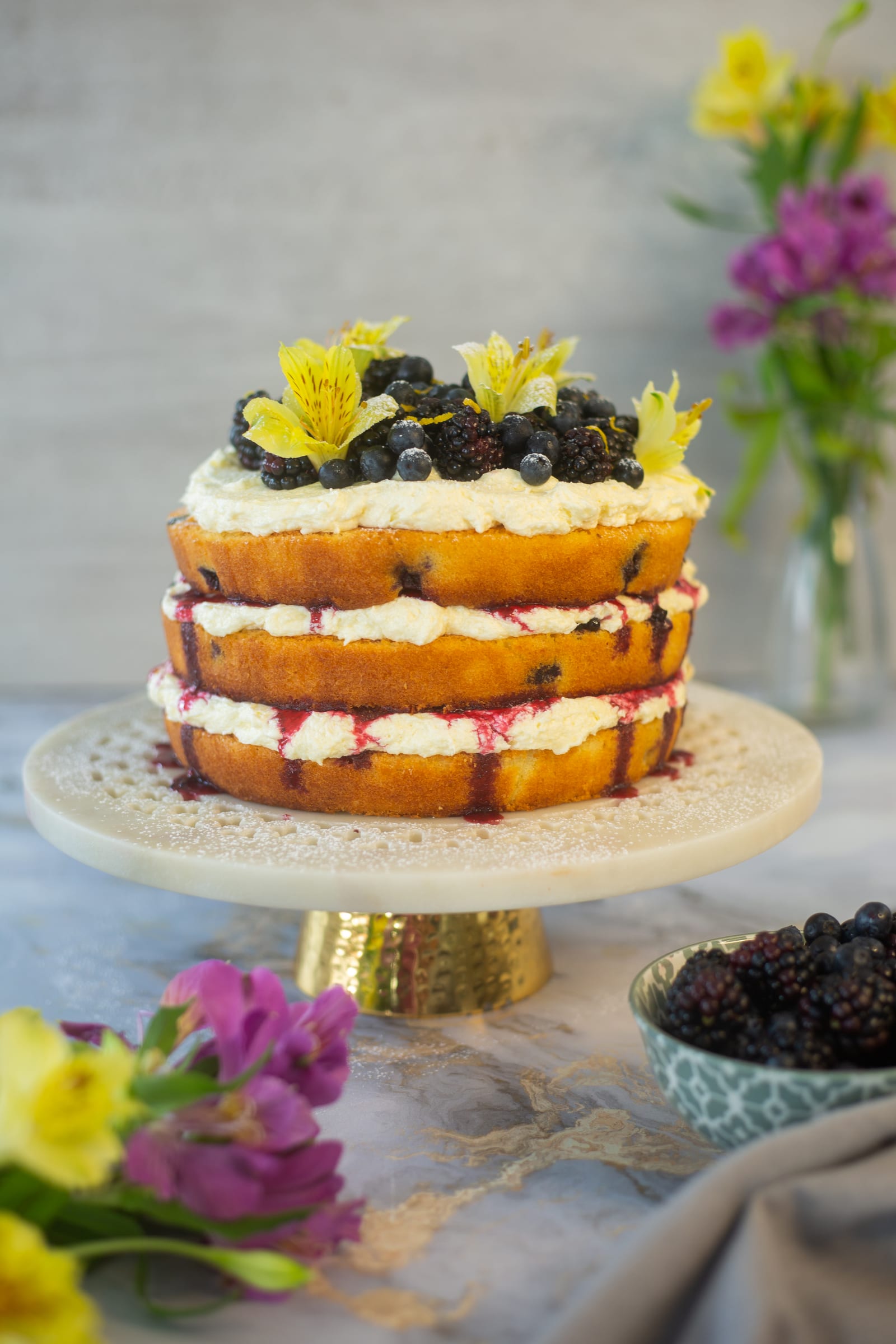 A whole lemon and blueberry cake on a cake stand, with purple flowers and a bowl of blackberries
