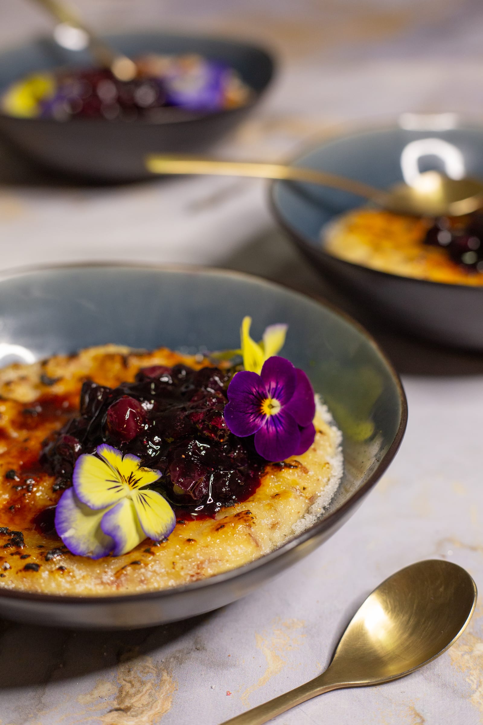 A bowl of kheer brûlée topped with blueberries and violas, with a gold spoon