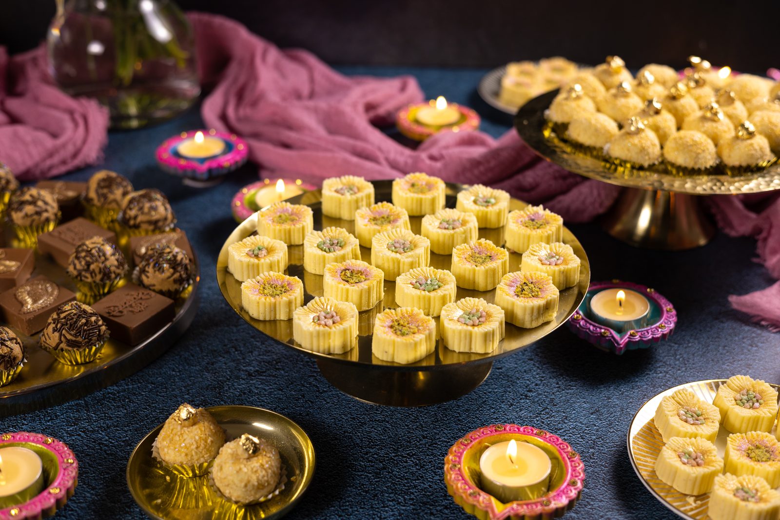 Flower-shaped simply wonderfuls on a gold cake stand, surrounded by lit diyas, flowers and other Diwali sweets