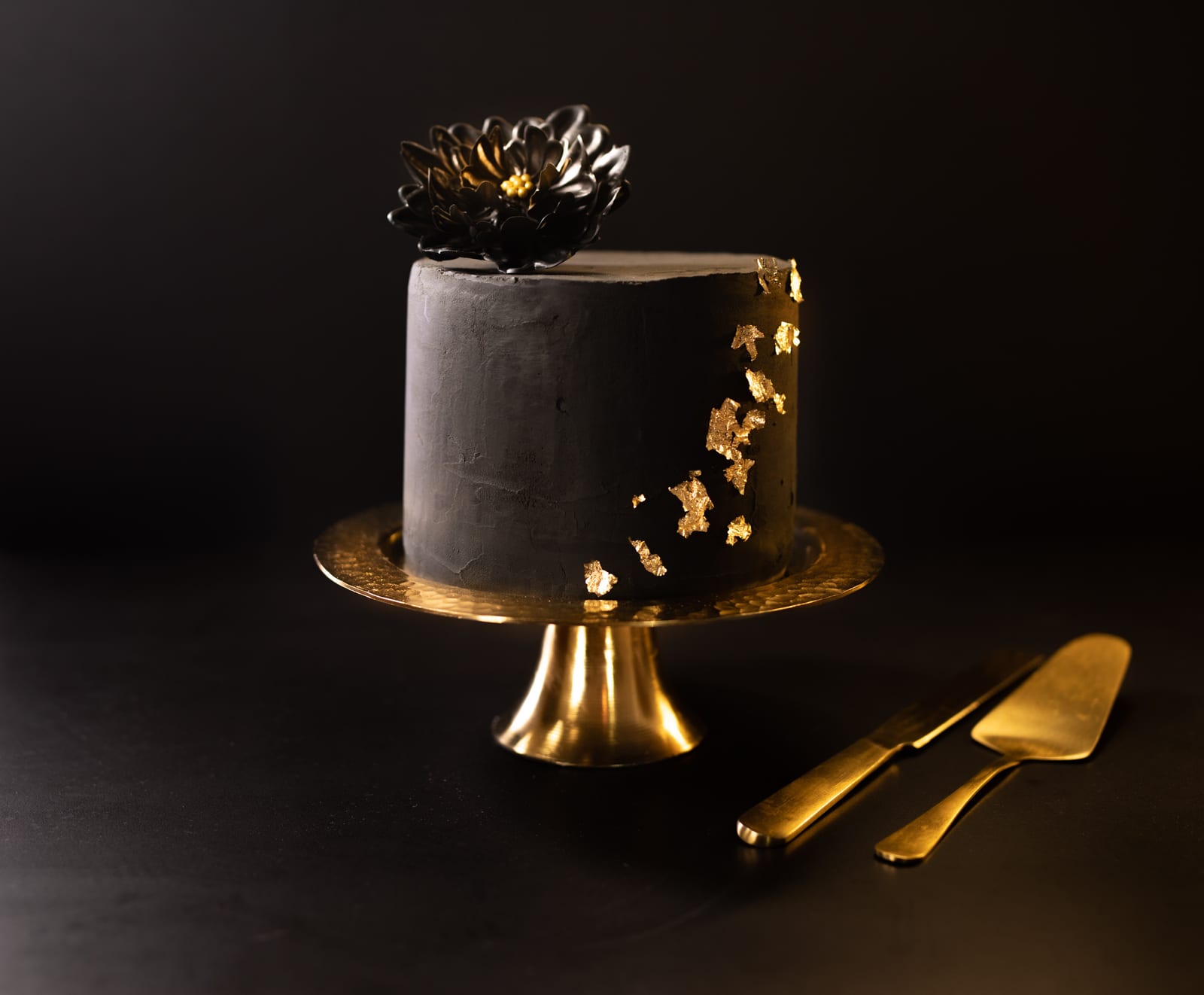 A matte black velvet cake with a black chocolate flower and gold leaf on a gold cake stand, a gold server beside it