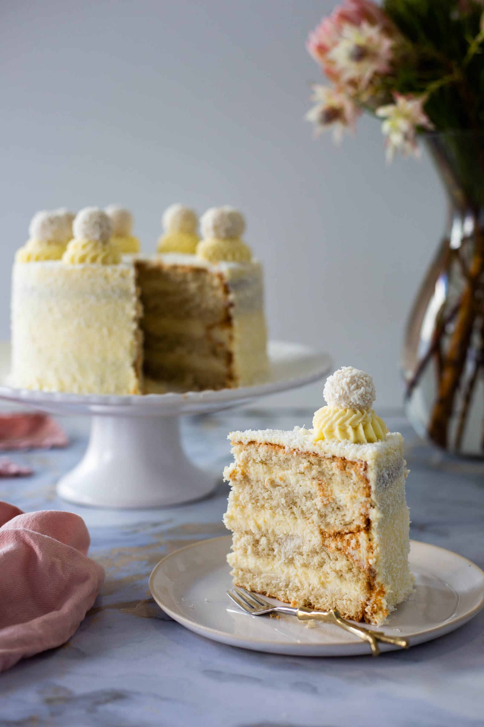 A slice of Raffaello cake in front of the cut cake on its stand, proteas behind